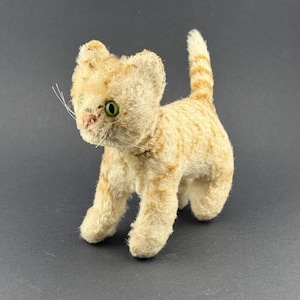 Könnte beinhalten: Ein Vintage-Stofftier-Katze in stehender Pose. Die Katze hat ein hellorangefarbenes und cremefarbenes Fell mit dunkleren orangefarbenen Streifen. Sie hat grüne Augen, eine rosa Nase und weiße Schnurrhaare. Der Schwanz der Katze ist aufrecht.