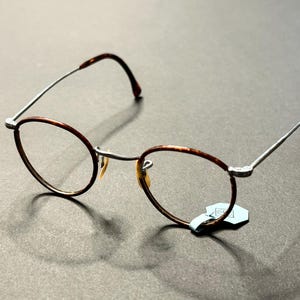 Peut inclure: Lunettes de forme ovale avec une monture marron à motif écaille de tortue sur les bords et les branches. Les montures sont en métal argenté. Les lunettes ont des verres transparents et une petite étiquette attachée à la monture.