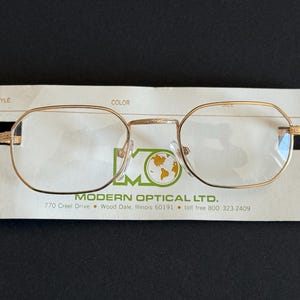 Op de afbeelding: Goudkleurige bril met een geometrische vorm, rustend op een witte kaart van Modern Optical Ltd. De kaart bevat het logo en adres van het bedrijf. De bril heeft heldere glazen en een klassiek ontwerp.