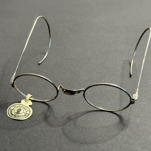 Peut inclure: Lunettes à monture ovale argentée avec des branches incurvées. Une petite étiquette avec du texte est suspendue à la monture. Les lunettes ont un design minimaliste, adapté aux accessoires vintage ou rétro.