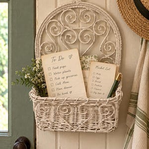 May include: A white wall-mounted wicker basket with a decorative arched metal back featuring scrollwork. The basket holds two paper cards with handwritten text: one titled To Do with a list of tasks, and one titled Market List with grocery items.