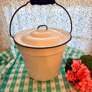 May include: Cream-colored enamelware bucket with a lid and a dark wooden handle. The bucket has a black rim and a wire handle. The background includes a floral patterned curtain and a green and white checkered tablecloth. A bouquet of pink flowers is on the right.