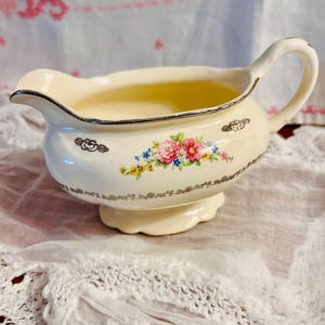 May include: Cream-colored vintage creamer with a silver trim and handle. The creamer features a floral design with pink, blue, and yellow flowers. The creamer sits on a small pedestal base. A decorative piece for serving cream or other liquids.