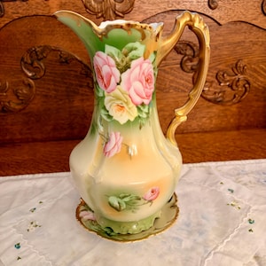 May include: A porcelain pitcher featuring a hand-painted floral design with pink and yellow roses on a cream and green gradient background. The pitcher has a scalloped rim, a gold-painted handle, and sits on a matching scalloped base.