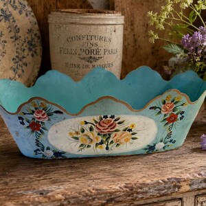 May include: An oval-shaped light blue metal bowl with a scalloped rim and a gold-toned edge. The exterior features a central white oval panel decorated with hand-painted pink, yellow, and red floral motifs, complemented by smaller floral accents.