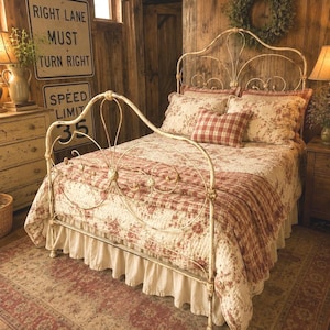 May include: A cream-colored metal bed frame with ornate scrollwork, dressed in a quilt featuring a red floral pattern and a red plaid throw. The bed includes two matching floral pillows and a small red plaid accent pillow, set atop a ruffled skirt.
