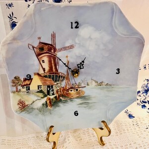 May include: A decorative clock with a Dutch windmill scene. The clock face is an irregular shape with a light blue background and black numerals. The clock is supported by a gold-colored metal stand.