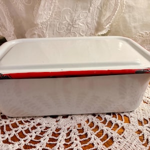 May include: A rectangular white enamelware bread pan with a lid. The pan has a red stripe around the rim and black trim. The pan is sitting on a white lace doily.