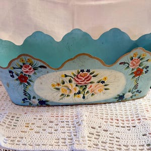 May include: A vintage, oval-shaped, light blue metal container with a scalloped edge. The container is adorned with hand-painted floral designs in pink, red, yellow, and blue. The rim is trimmed with gold paint. A decorative piece.