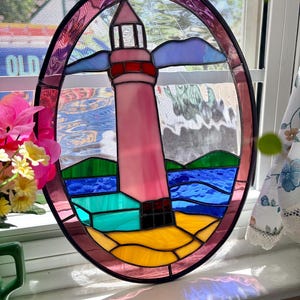May include: Oval stained glass window art featuring a pink lighthouse with a red top, blue sky, and green and blue water. The lighthouse sits on a yellow and teal beach. The frame is a dark purple color.