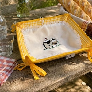 May include: A square fabric bread basket featuring a yellow and white gingham border, white lace trim, and a central cream-colored panel embroidered with a cow and chicken motif. The corners are secured with yellow fabric ties and a Leonardo label.