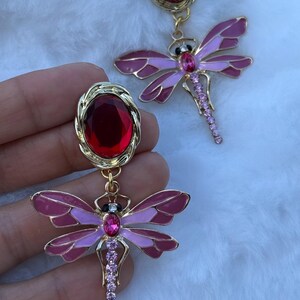 Puede incluir: Un par de pendientes dorados con una gran piedra preciosa de color rojo oscuro de corte ovalado engastada en un marco de metal trenzado. De cada piedra cuelga un dije de libélula con alas de esmalte rosa y una cola con cristales rosados.