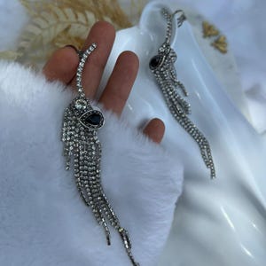 Puede incluir: Un par de pendientes colgantes plateados. Cada pendiente presenta una parte superior curvada con una piedra transparente, una piedra negra en forma de lágrima y múltiples hebras de pequeñas piedras claras brillantes. Los pendientes están diseñados para captar la luz.