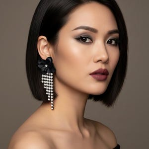 Puede incluir: Una mujer con cabello oscuro y maquillaje lleva un pendiente de flor negra con brillantes en cascada. El pendiente está unido a una flor negra. La mujer lleva un top negro con los hombros descubiertos.