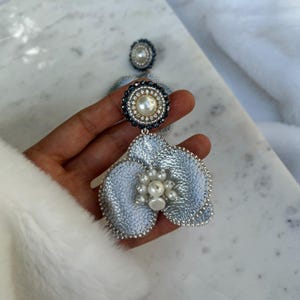 Puede incluir: Un par de pendientes en forma de flor azul claro con detalles de perlas y cristales. Los pendientes tienen un borde plateado y un centro de perlas. La parte superior de los pendientes tiene un perno de perla y cristal.