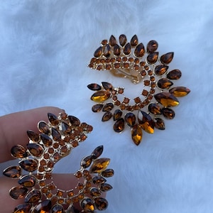 May include: A pair of crescent-shaped clip-on earrings featuring amber-colored, teardrop-shaped crystals arranged in a sunburst pattern. The metal base is gold-toned, and the design is accented with smaller, round, amber-colored rhinestones.