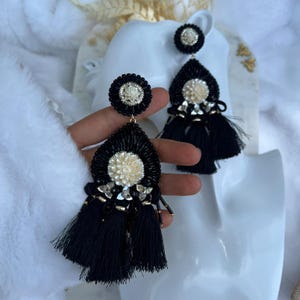 Puede incluir: Pendientes negros con cuentas en forma de lágrima, con un diseño de flor color crema y borlas negras. Los pendientes tienen un perno circular con un diseño de flor a juego. Los pendientes se exhiben en una cabeza de maniquí blanca.