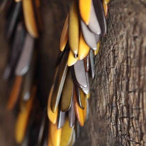 Op de afbeelding: Een close-up van een lange, vallende ketting gemaakt van kleine, druppelvormige stukjes hout. Het hout is een mix van bruine, grijze en gele tinten. De ketting hangt tegen een ruwe, houten achtergrond.
