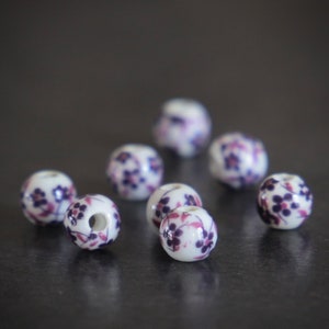 Könnte beinhalten: Eine Gruppe von acht weißen Keramikperlen mit einem violetten Blumenmuster. Die Perlen sind rund und haben ein Loch in der Mitte.