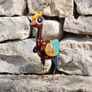 Op de afbeelding: Een kleurrijk metalen vogelsculptuur met een gele kroon, bruin lichaam, blauwe en gele vleugels en grote witte ogen. Het beeld staat op een stenen oppervlak.