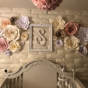 Puede incluir: Una cuna blanca con un marco blanco y un dosel blanco. La pared detrás de la cuna está decorada con flores de papel en tonos rosa, blanco y marrón. Las flores están dispuestas en un círculo alrededor de un marco blanco con la letra "S" en el centro.
