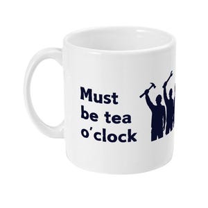May include: A white ceramic mug featuring dark blue text that reads Must be tea o'clock. Next to the text is a dark blue silhouette graphic of three figures holding tools, including a hammer and a wrench, standing in a row.