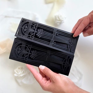 May include: Black laser cut wedding invitation with an intricate geometric design. The invitation is folded in half and held by a hand.