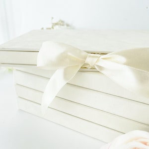 May include: A stack of four rectangular, off-white boxes with smooth, matte surfaces. The top box is secured with a wide, shiny, cream-colored satin ribbon tied into a bow. A single, pale pink and white artificial flower rests near the base.