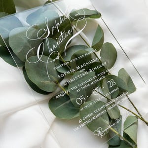 May include: A clear acrylic wedding invitation with elegant white script. The invitation includes names, date, time, and location details. Green eucalyptus leaves are arranged behind the invitation, adding a natural touch.