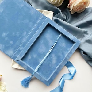 Puede incluir: Una caja rectangular azul con acabado texturizado, que cuenta con una tapa abatible y un marcapáginas de cinta azul a juego con una borla. El interior de la caja está abierto, revelando un compartimento profundo y vacío.