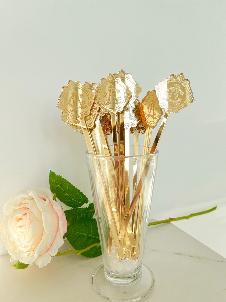 May include: A set of gold-colored metal stirrers with decorative tops in a glass vase. The tops feature a floral design with a monogram.