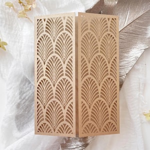 May include: A gold-colored, Art Deco-style invitation card. The card is folded and features a repeating fan-shaped pattern cut into the surface. The card is displayed on a white and silver background.