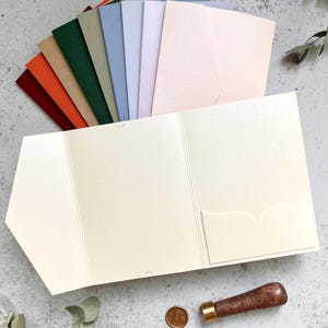 May include: An assortment of colorful stationery items, including envelopes and cards, arranged on a textured surface. The color palette includes shades of red, orange, green, blue, and pink. A cream-colored pocket folder, a wax seal, and a wooden handled stamp are also present.