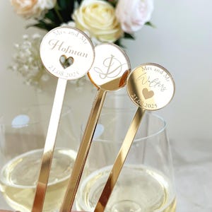 May include: Three gold-toned mirrored acrylic drink stirrers with circular tops. Two stirrers feature the text "Mrs and Mr" followed by a surname, a heart cutout, and the date "21.08.2025". The center stirrer displays a large, elegant script letter "D".