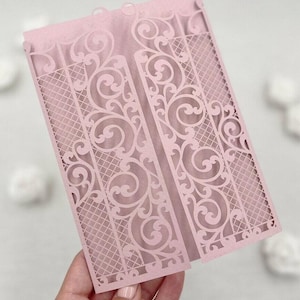 May include: A light pink laser-cut invitation card with an ornate gate design. The card features intricate scrollwork and a lattice pattern, creating a delicate and elegant aesthetic. The card is folded and held open, showcasing the detailed design.