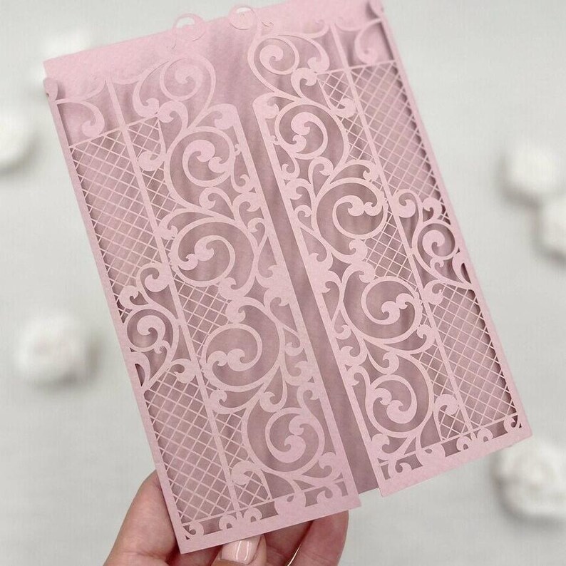 May include: A light pink laser-cut invitation card with an ornate gate design. The card features intricate scrollwork and a lattice pattern, creating a delicate and elegant aesthetic. The card is folded and held open, showcasing the detailed design.
