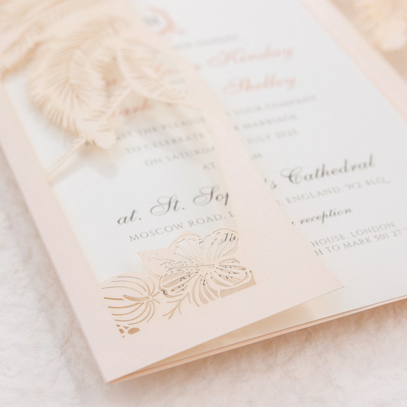 May include: A light peach-colored wedding invitation with a floral cut-out design. The invitation is open, revealing printed text. The design includes a decorative feather element and the text "at St. Sop" and "Moscow Road, London".