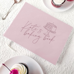 May include: A pink baking book with the text "Katie's baking book" in cursive script. A line drawing of a cake on a cake stand is next to the text. Two cupcakes with white frosting and cherries are in the background.