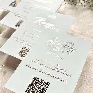 Puede incluir: Tarjetas de respuesta rectangulares blancas con letras en l&aacute;mina dorada que dicen Kindly Reply by 17.08.29. Cada tarjeta incluye un c&oacute;digo QR impreso, instrucciones para escanear el sitio web de la boda y la URL www.angelinaethanwedding.com en negro.