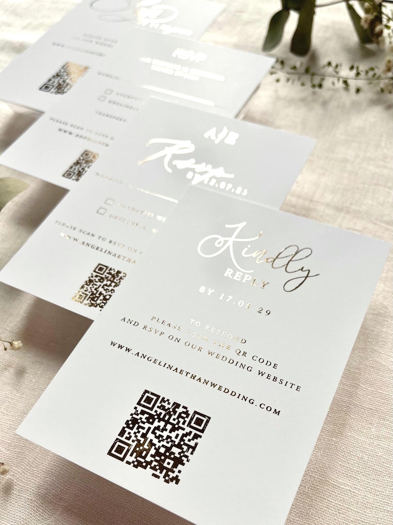 Puede incluir: Tarjetas de respuesta rectangulares blancas con letras en l&aacute;mina dorada que dicen Kindly Reply by 17.08.29. Cada tarjeta incluye un c&oacute;digo QR impreso, instrucciones para escanear el sitio web de la boda y la URL www.angelinaethanwedding.com en negro.