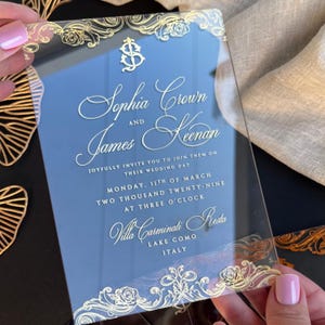 May include: Clear acrylic wedding invitation with gold floral accents and elegant script. The invitation reads "Sophia Crown and James Keenan" and includes the wedding date, time, and location in Lake Como, Italy.