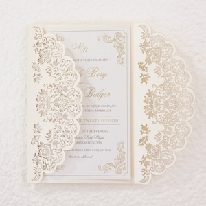Peut inclure: Une invitation de mariage blanche avec un motif découpé au laser de couleur or. L'invitation comprend les noms "ND", "Berg" et "Bulger", la date "April Twenty Seventh" et le lieu "Veston Park Plaza, Massachusetts".
