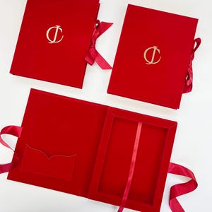 Puede incluir: Tres cajas rectangulares de terciopelo rojo con cierres de cinta. Dos cajas están cerradas, cada una con un emblema dorado de la letra D en la tapa. Una caja está abierta, revelando un interior rojo con un bolsillo y una cinta.