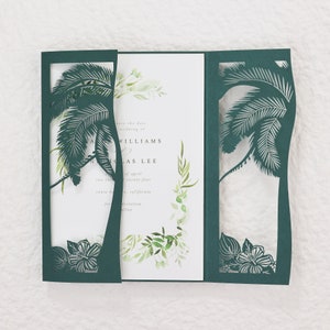 May include: A dark teal wedding invitation with a laser-cut palm tree design. The invitation has a tri-fold design with the text "save the date" and names. The inner panel features watercolor greenery.