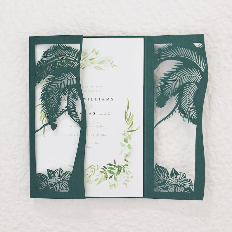 May include: A dark teal wedding invitation with a laser-cut palm tree design. The invitation has a tri-fold design with the text "save the date" and names. The inner panel features watercolor greenery.