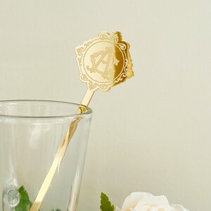 May include: Gold-toned metal stirring stick with a decorative, ornate, square-shaped top featuring the letter "A" in a monogram design. The stick is in a clear glass cup.