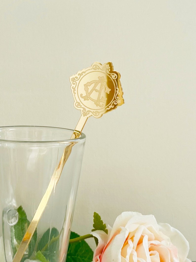 May include: Gold-toned metal stirring stick with a decorative, ornate, square-shaped top featuring the letter "A" in a monogram design. The stick is in a clear glass cup.