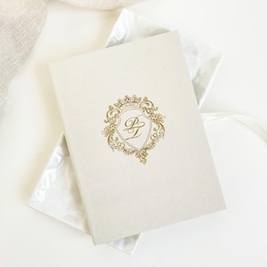 May include: Cream-colored wedding invitation with a gold crest design. The crest features the initials "PL" and the date "1.10.2027". The invitation is presented in a white box with a decorative pattern.