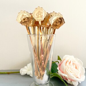 May include: A set of gold-colored cocktail stirrers with a decorative monogram design. The stirrers are in a clear glass vase with a white cotton ball at the bottom. A pink rose is in the background.
