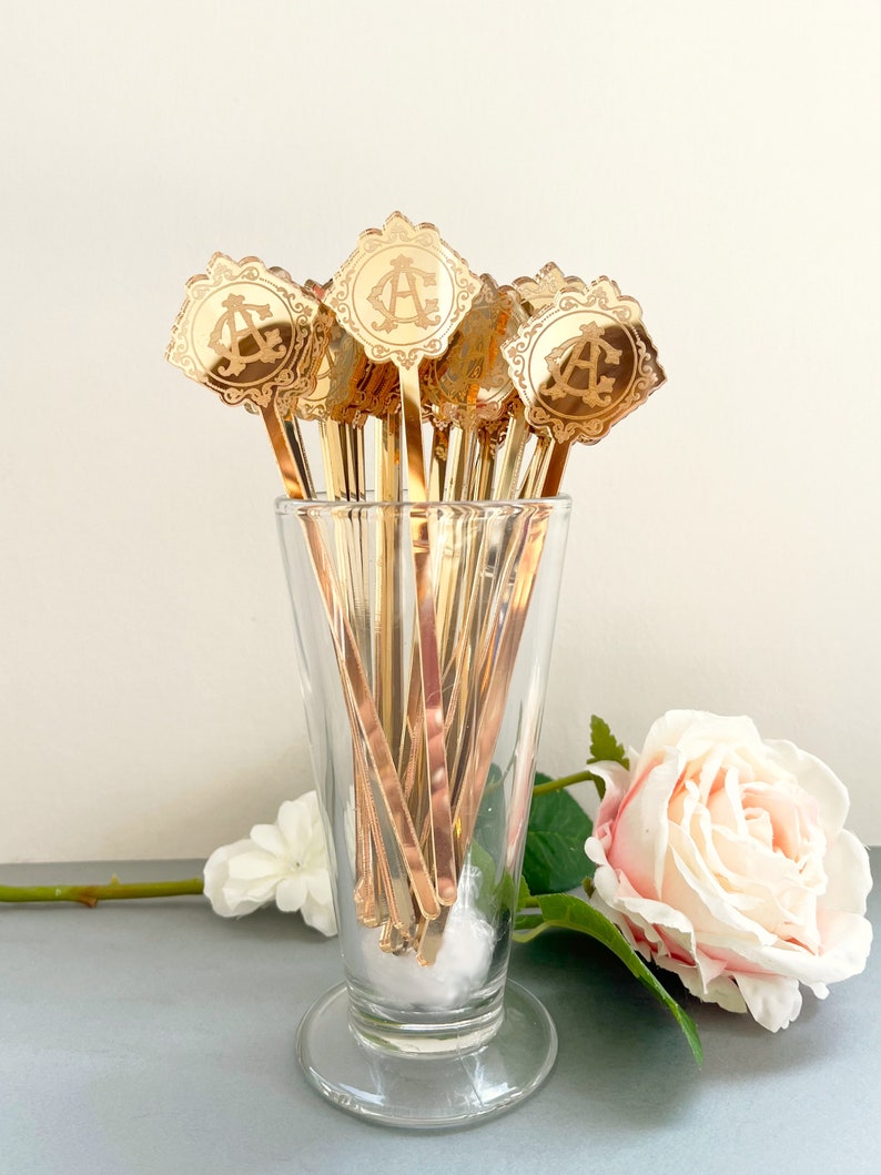 May include: A set of gold-colored cocktail stirrers with a decorative monogram design. The stirrers are in a clear glass vase with a white cotton ball at the bottom. A pink rose is in the background.